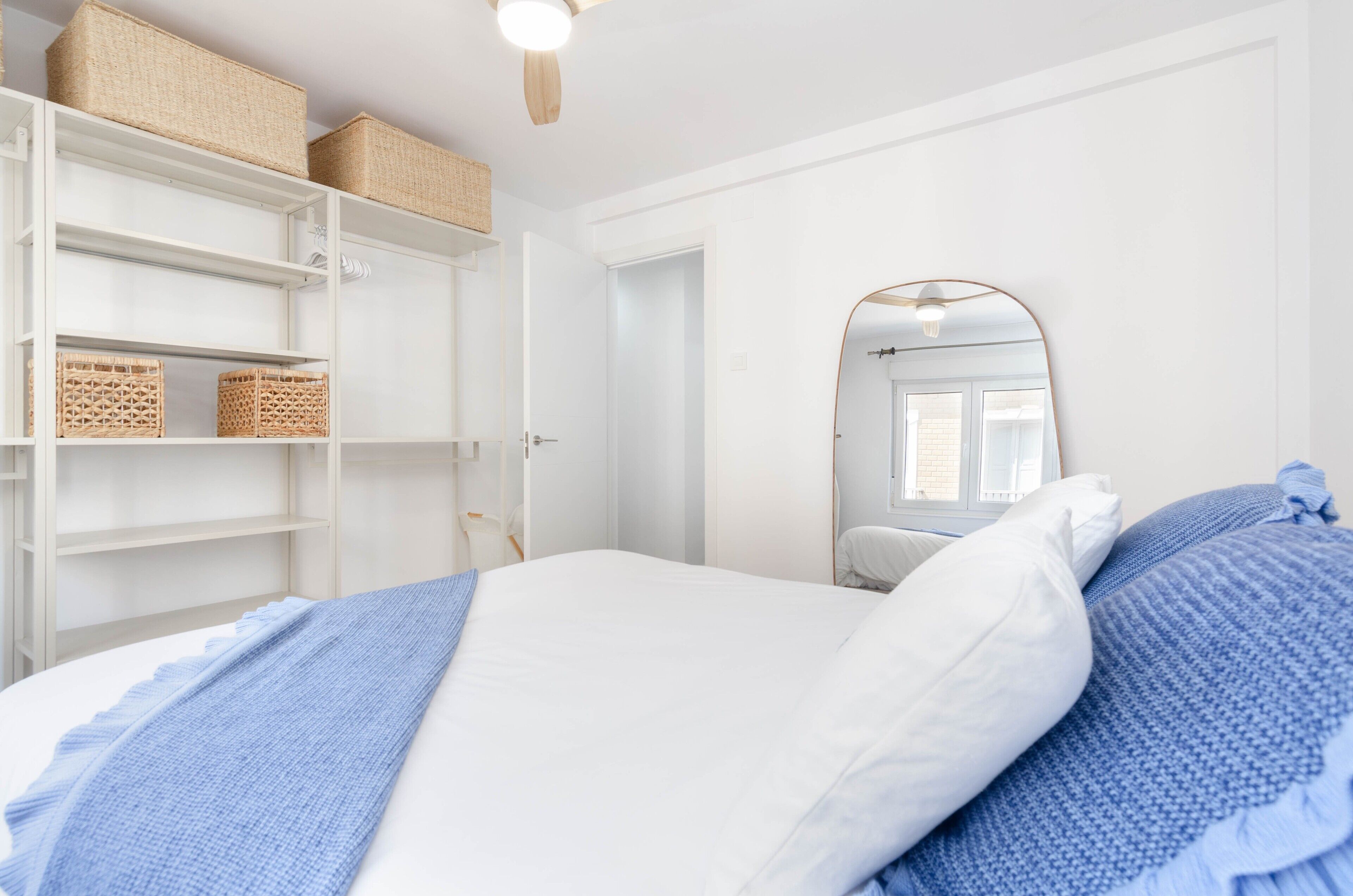 A bedroom with a bed, white walls, a wooden ceiling, a wardrobe with baskets, and a mirror.