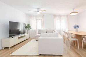 A modern living room with a flat-screen TV, a sofa, a dining table with chairs, and a ceiling fan.
