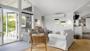 Un vaste espace de vie avec une grande fenêtre, un canapé blanc, un fauteuil en osier et un coin cuisine visible à travers une porte ouverte.