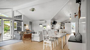Une cuisine spacieuse avec des meubles blancs, une table à manger et des chaises. Une grande fenêtre laisse entrer la lumière naturelle et offre une vue sur l’extérieur.