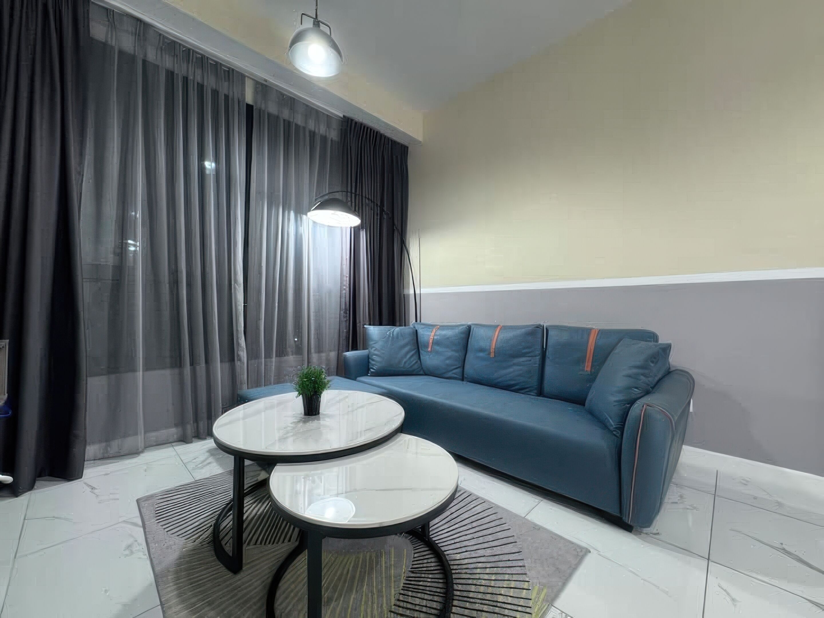 Una sala de estar moderna con un sofá azul, dos mesas redondas, una planta en maceta y un ventanal con cortinas traslúcidas.