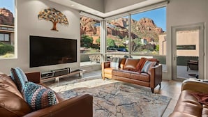 A spacious living room with a large flat-screen TV, leather sofas, a patterned rug, and a mountain view through large windows.