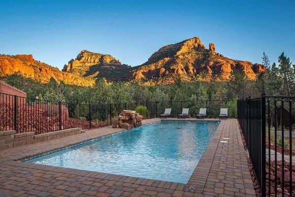 A rectangular pool with a stone waterfall feature, surrounded by a brick patio and black metal fencing.