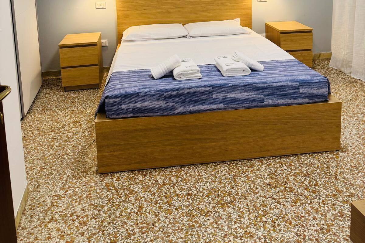 A neatly made bed with white linens and a blue patterned bedspread, flanked by wooden nightstands with lamps, in a room with a speckled floor and light blue walls.