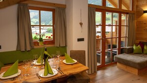 A dining area with a wooden table set for four, a sofa, and a view of a mountainous landscape through large windows. - Apartment 'Deluxe 2' with Private Garden, Balcony and Wi-Fi (Großarl)