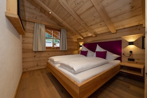 Un dormitorio de cabaña de madera con cama, una ventana con cortinas, una mesita de noche y una lámpara fijada a la pared.