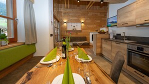A dining area with a wooden table set for two, a kitchen with wooden cabinets, and a television mounted above the counter. - Apartment 'Deluxe 2' with Private Garden, Balcony and Wi-Fi (Großarl)