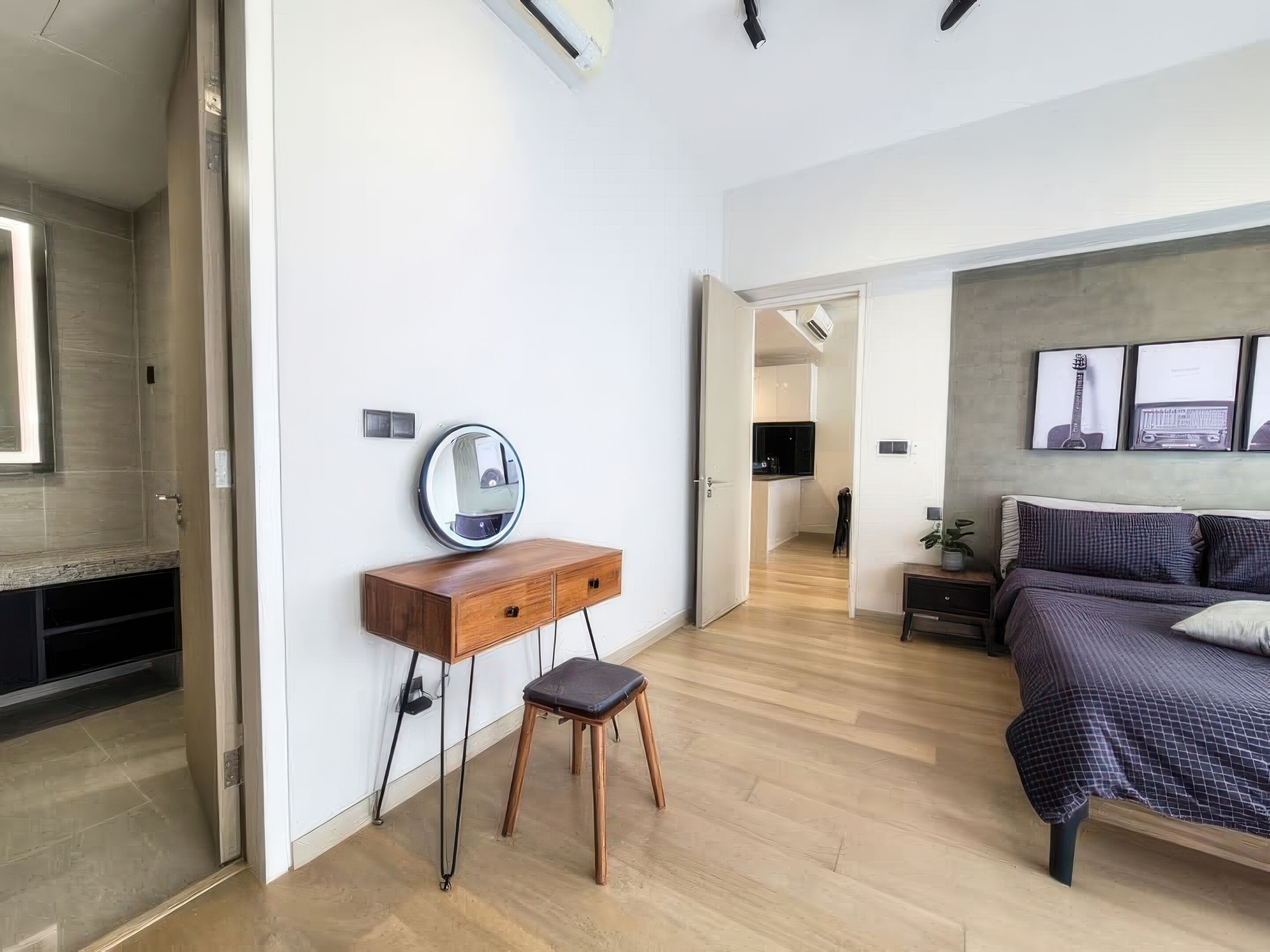 A modern bedroom with a wooden dresser, a round mirror, a bed with a checkered duvet, and a small stool.