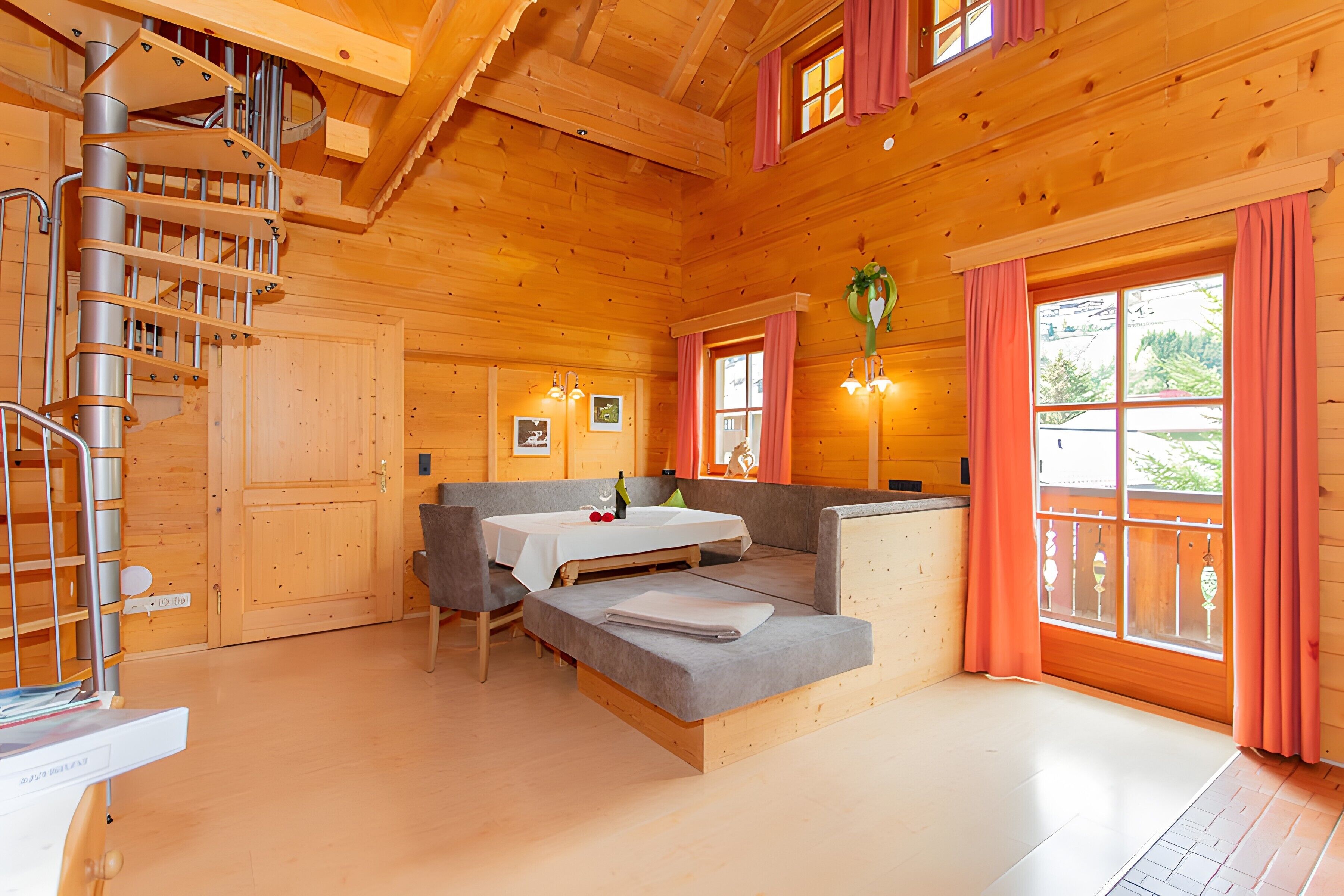 Een houten chalet interieur met een spiltrap, een slaapbank, een eettafel en een raam met gordijnen.