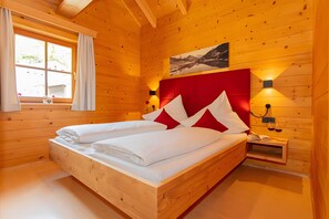 Een houten kamer in chaletstijl met een tweepersoonsbed, een raam met gordijnen, een hangend schilderij en bedlampjes.