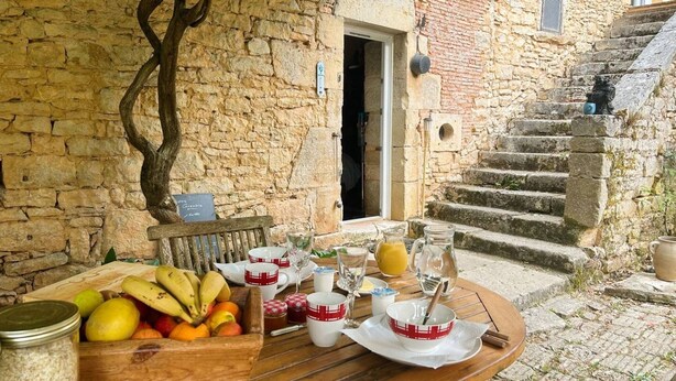 A breakfast setup with fruits, cups, and a jug on a wooden table outside a stone building with a staircase.