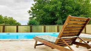 A wooden lounge chair with a patterned cushion by a swimming pool, surrounded by a wooden fence and lush greenery.