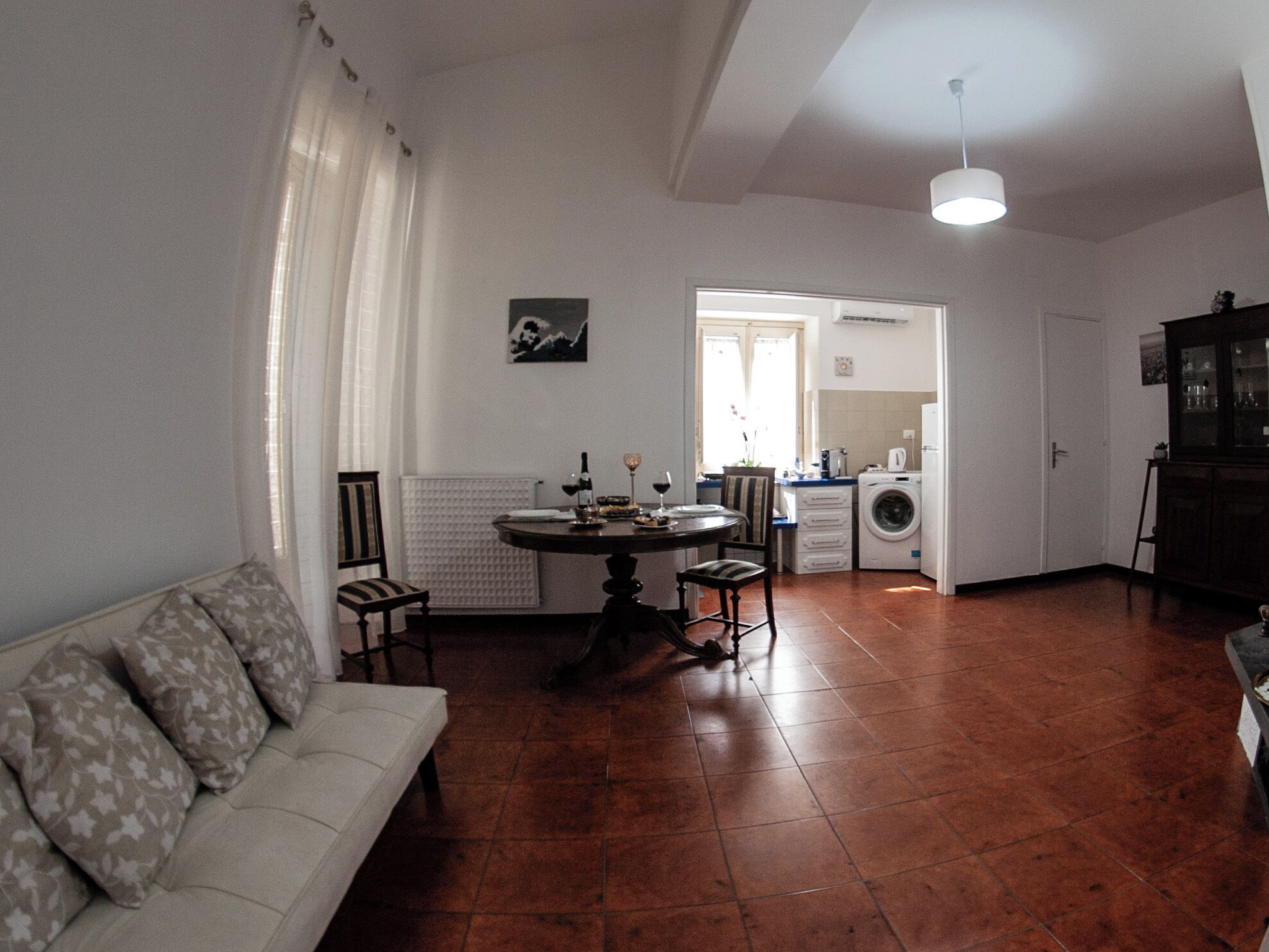 A living room with a sofa, a dining table, and chairs. There is a kitchen area with a washing machine and a cabinet.