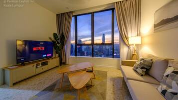 A modern living room with a large TV, a sofa, a wooden table, and a view of the cityscape through large windows.