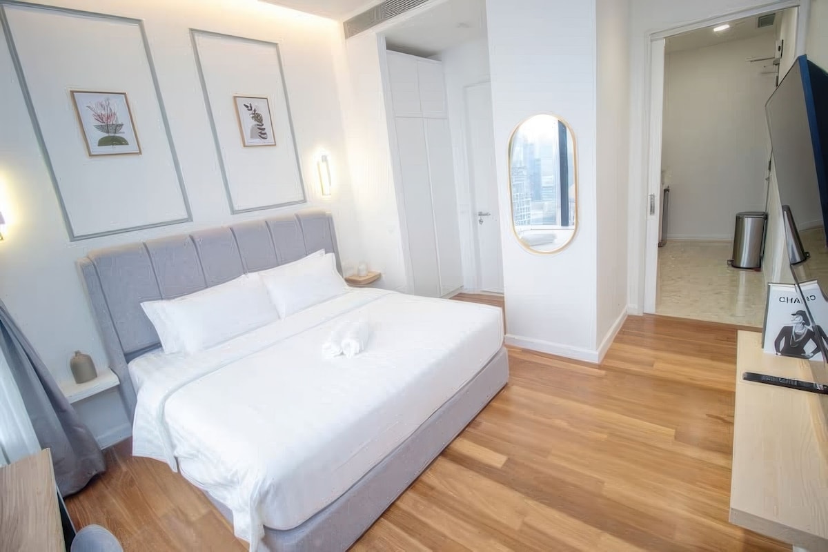 A modern hotel room with a large bed, a bedside table, a mirror, and framed artwork on the wall.