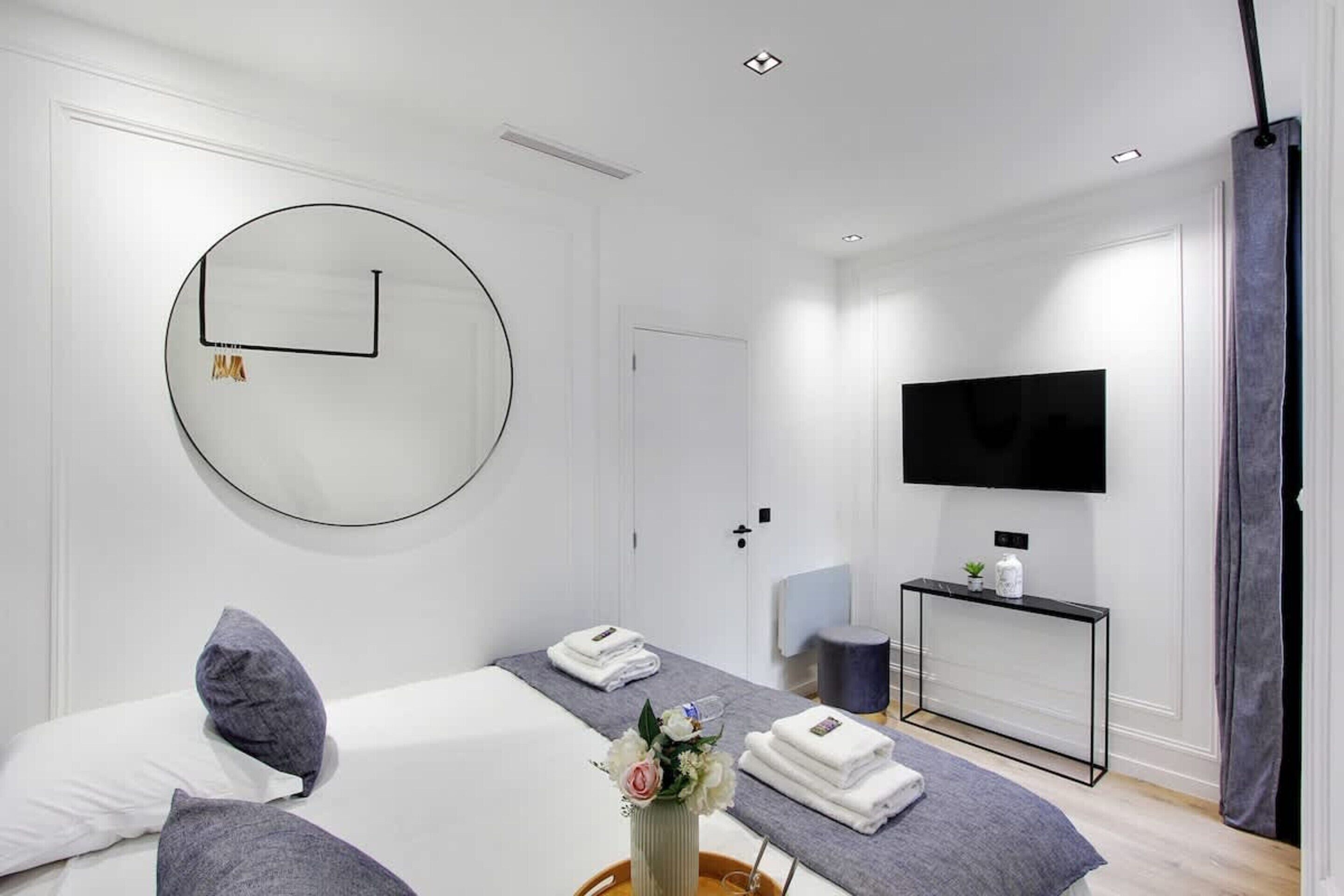 A modern hotel room with a large round mirror, a flat-screen TV, a bed with a grey headboard, a bedside table with a vase of flowers, and a small console table with a TV and a plant.