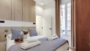 Une chambre d’hôtel avec un lit, des oreillers, un plateau avec des fruits et des fleurs, et une vue sur l’extérieur.