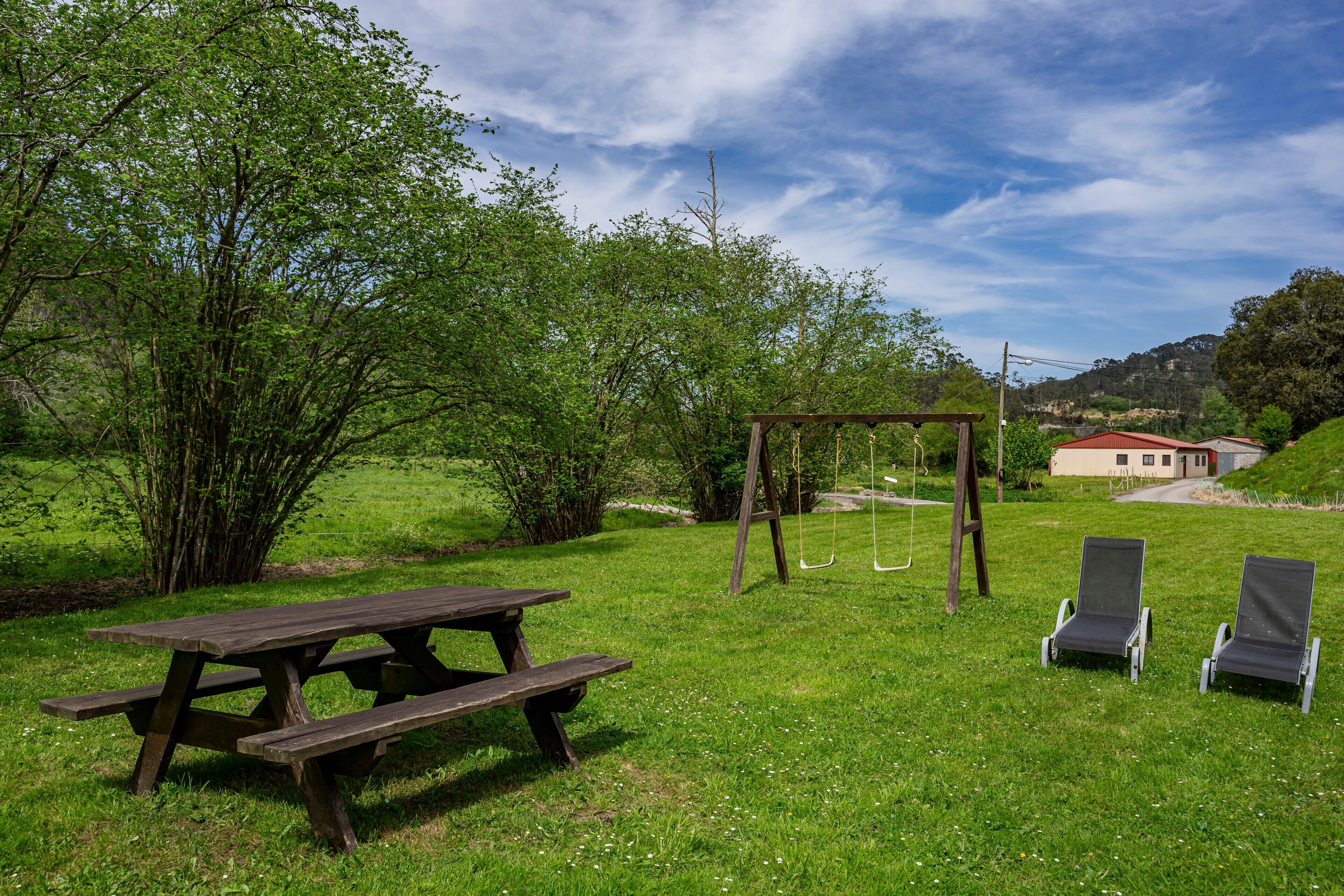 A picnic table, a swing set, and two lounge chairs in a grassy area with trees and a building in the background.