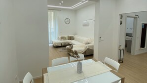 Un espace repas moderne avec une table et des chaises blanches, un vase contenant des plantes séchées et une vue sur un salon avec un canapé et une lampe.