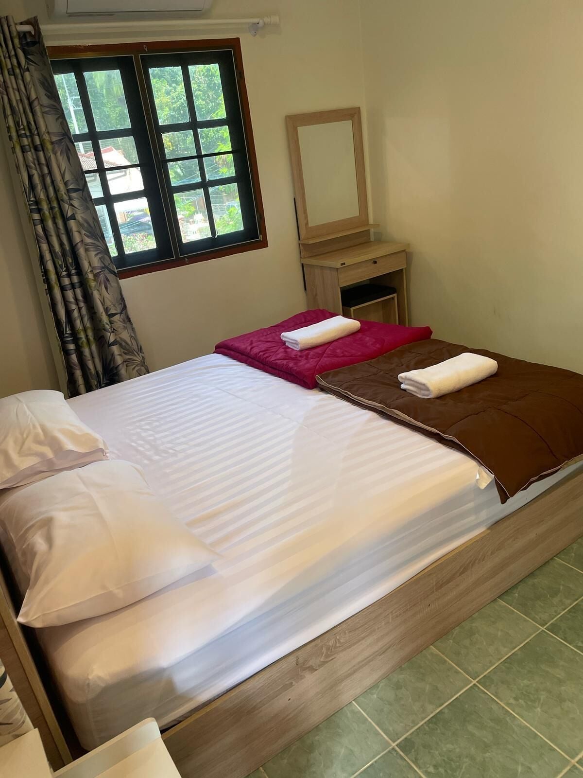 A double bed with white and brown bedding, a wooden nightstand with a mirror, and a window with curtains.