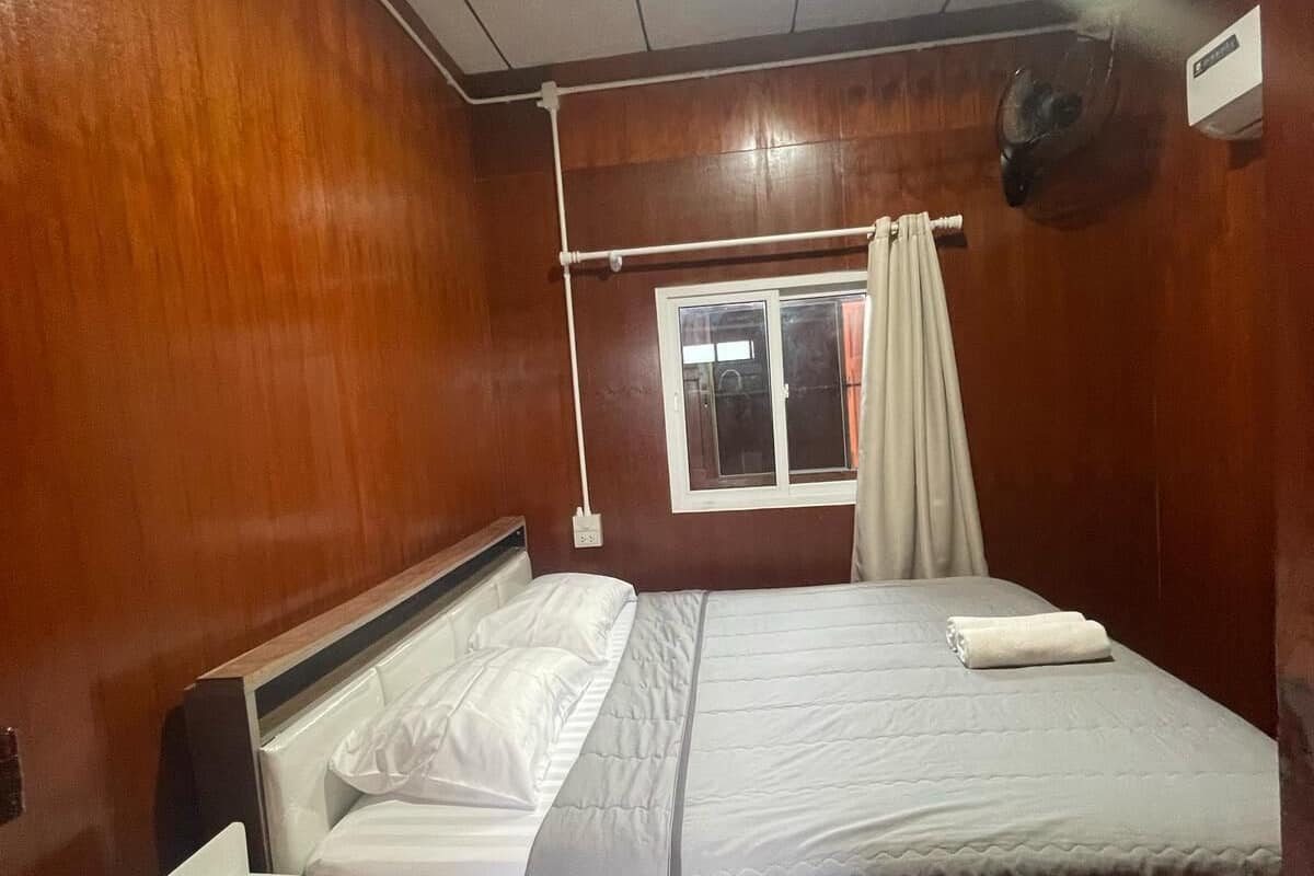 Habitación de hotel compacta con interior de madera, una cama individual con ropa blanca, una mesita de noche y una ventana con cortinas.