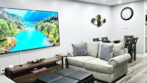Ein modernes Wohnzimmer mit Flachbildfernseher, grauem Sofa, schwarzem Hocker und Couchtisch aus Holz.