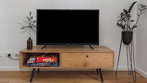 Un mueble de televisión de madera con un televisor de pantalla plana, un estante pequeño con una caja de juegos y una planta en maceta.