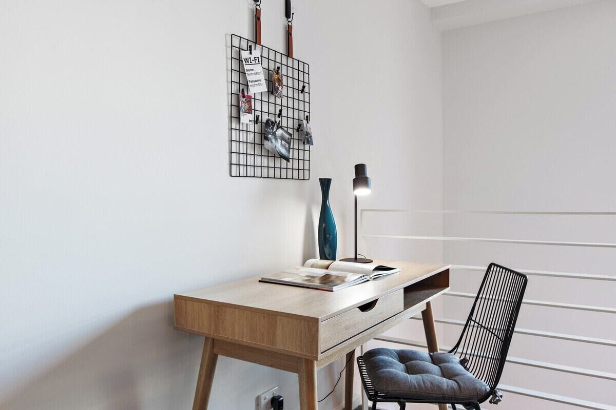 A modern desk with a light wood finish, a black chair with a cushioned seat, and a wall-mounted organizer with various items.