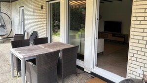 Eine Terrasse mit Tisch und Stühlen, einem Schaukelstuhl und einer Glastür, die zu einem Innenbereich mit Fernseher und Holzschrank führt.