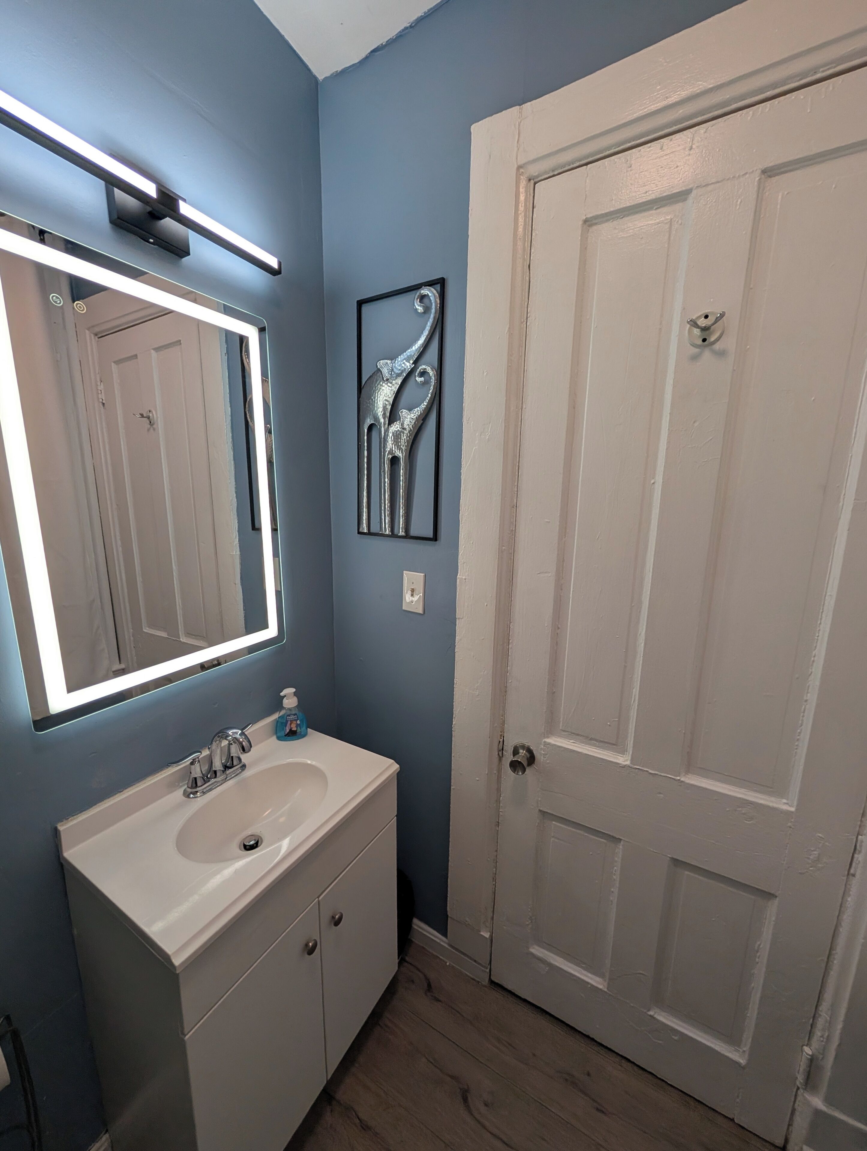 A bathroom with a white sink, a mirror with underglow lighting, a door, and a wall-mounted light fixture.