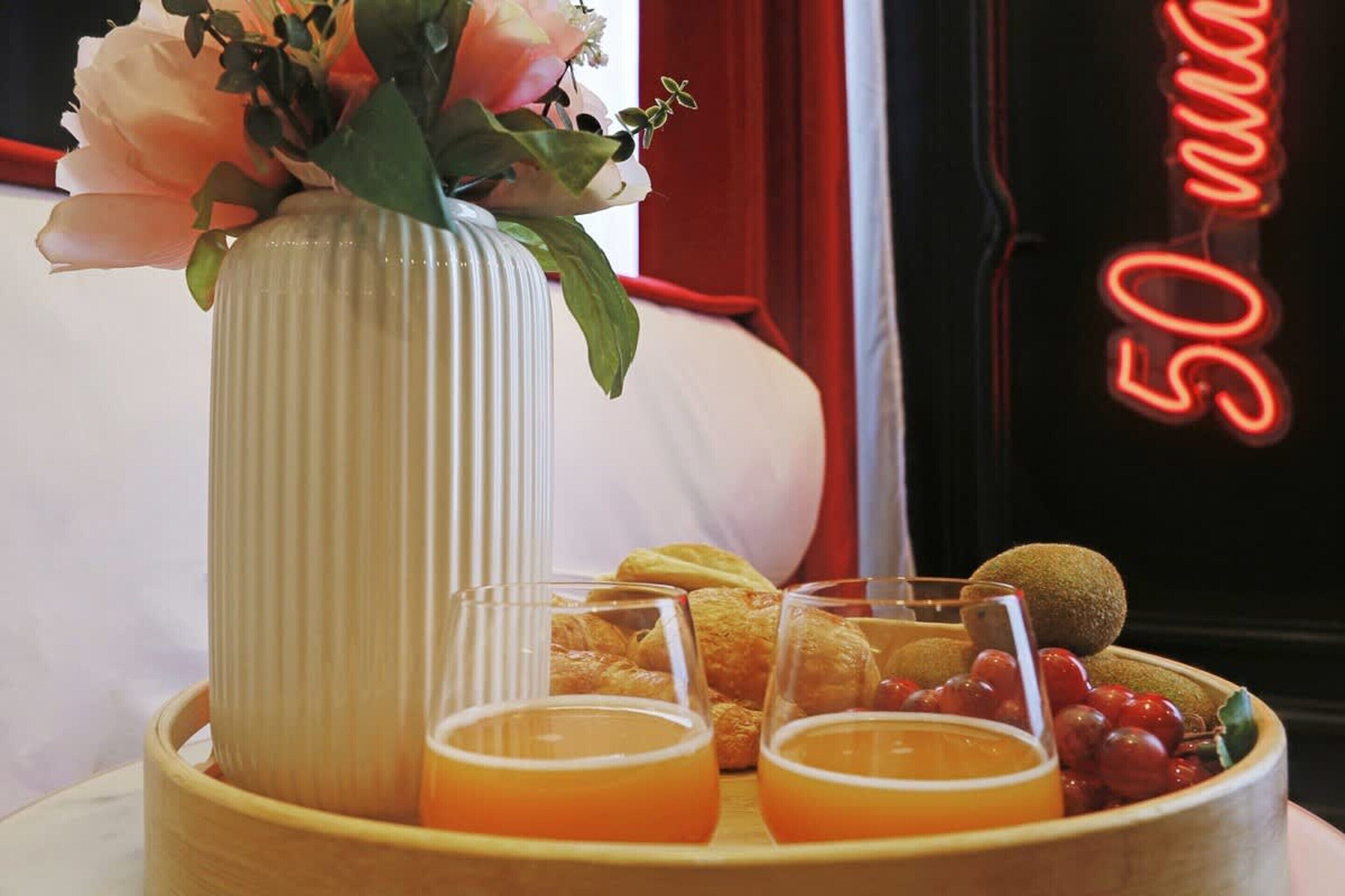 Uma bandeja de café da manhã com croissants, kiwi, uvas e copos de suco de laranja, um vaso com flores e um letreiro de neon ao fundo.
