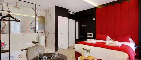 A hotel room with a bed, a dining table with chairs, a mirror, and a small side table with a vase and a tray of food.