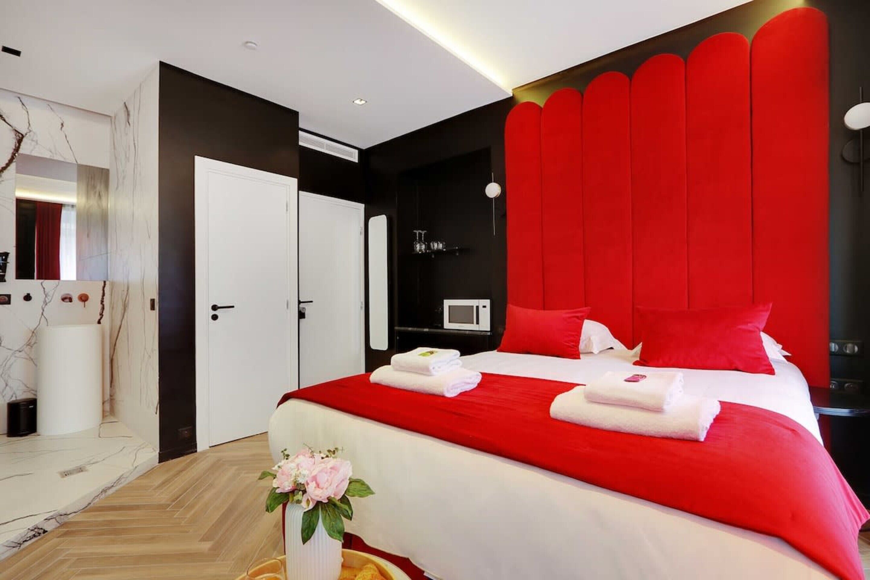 A modern hotel room with a large bed, red accent walls, a white door, and a wooden floor.