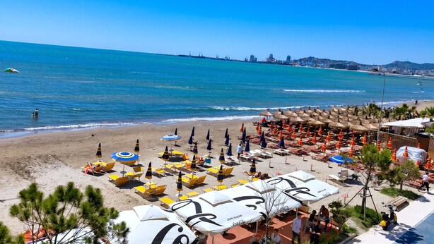 Una playa con sombrillas y sillas reclinables, cielo azul despejado y un horizonte con edificios.