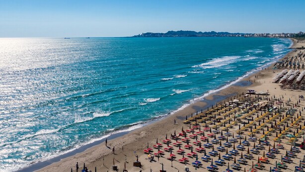 A beach with rows of umbrellas and sun loungers, a clear blue sea, and a distant city skyline.