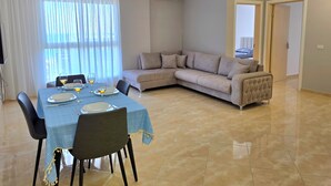 Una sala moderna con un sofá modular, una mesa de comedor para dos personas y un ventanal con cortinas translúcidas.