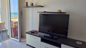 A modern living room with a flat-screen TV on a dark console, a glass door leading to a balcony with a view of the beach, and a small shelf with decorative items. - Beachfront & Sea View Apartment 102 (Durres)