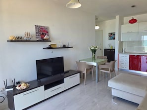 Een moderne woonkamer met een flatscreen televisie, een eettafel, stoelen en een keukenhoek met kasten en een rode apparatuur.
