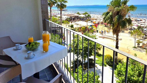 A balcony with a table set for breakfast, overlooking a beach with umbrellas and palm trees. - Beachfront & Sea View Apartment 102 (Durres)