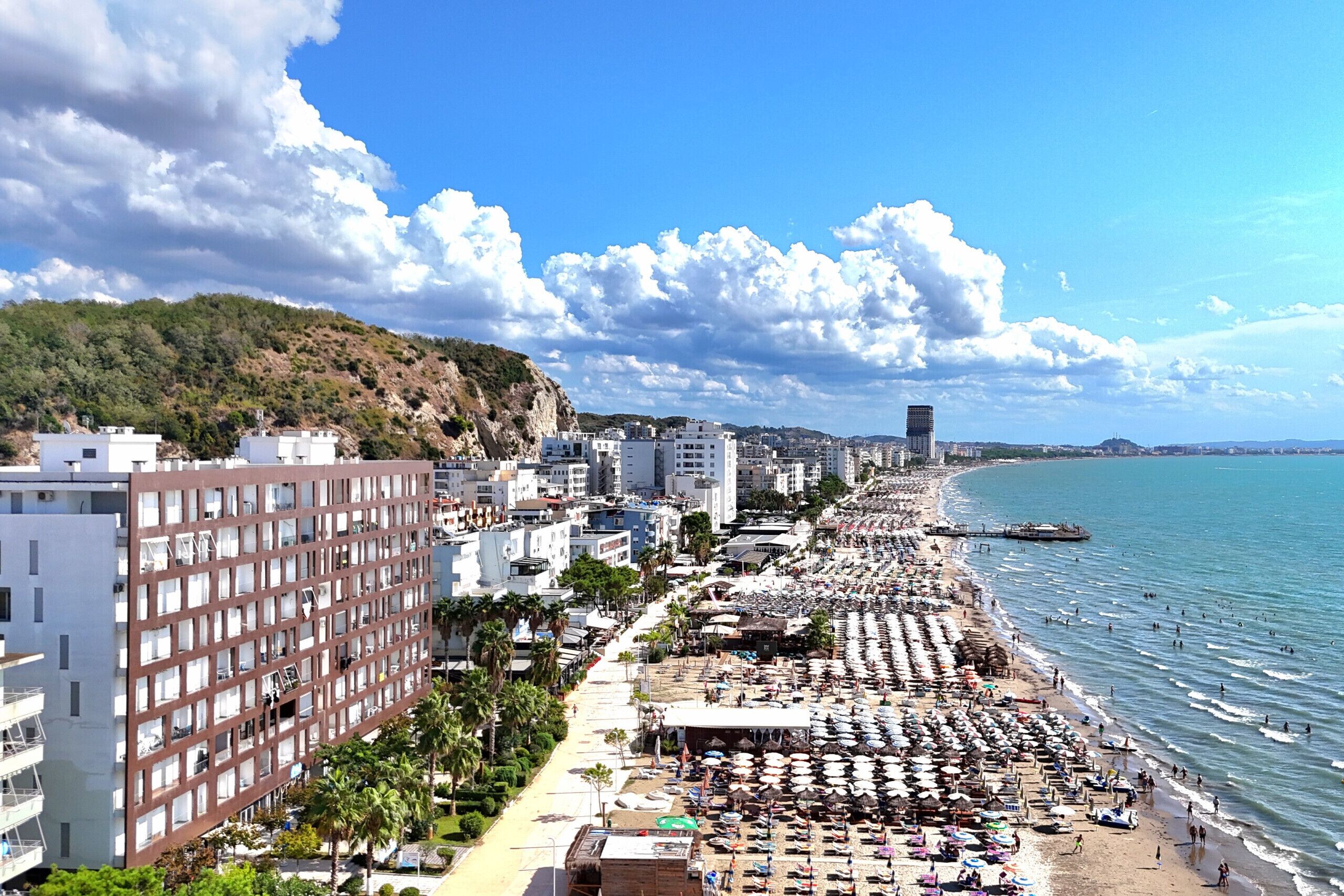 A coastal cityscape with a long beach lined with umbrellas and sun loungers, buildings, and a clear sky.