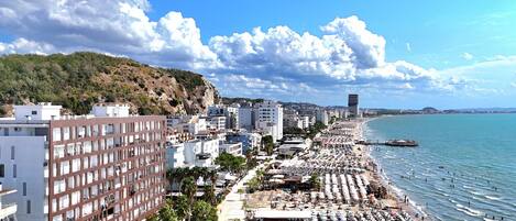A coastal cityscape with a long beach lined with umbrellas and sun loungers, buildings, and a clear sky.