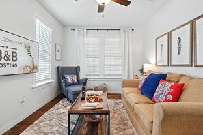 A living room with a sofa, a coffee table, a chair, and a ceiling fan. There are framed pictures on the wall and a window with blinds.
