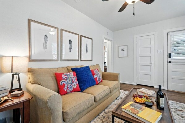 A living room with a beige sofa, a coffee table with books and a bottle, and framed artwork on the wall.