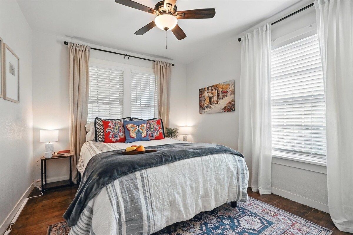 Une chambre à coucher avec un lit, un ventilateur de plafond, une table de chevet, une lampe et une fenêtre avec des stores.