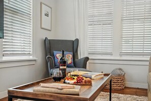 A cozy living room with a grey armchair, a wooden coffee table with a bottle, glasses, and a fruit platter.