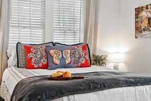 A bed with patterned pillows, a bedside table with a lamp, and a window with blinds.