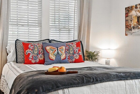A bed with patterned pillows, a bedside table with a lamp, and a window with blinds.