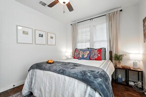 A bedroom with a bed, a ceiling fan, a window with curtains, and framed pictures on the wall.