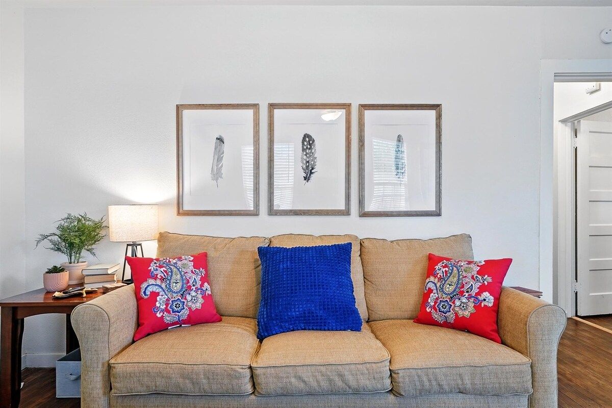 Un salon avec un canapé beige, deux coussins rouges à motifs, un coussin bleu et trois œuvres d’art encadrées représentant des plumes, accrochées au mur.