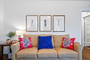 A living room with a beige sofa, two red patterned pillows, a blue pillow, and three framed feather artworks on the wall.
